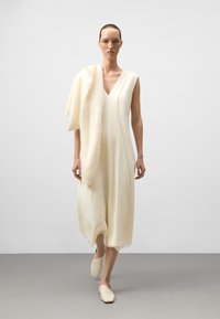 Cremefarbenes ärmelloses Kleid mit V-Ausschnitt, fließendem Design, geschichteter Textur und einem weichen Schal, der über eine Schulter drapiert ist, kombiniert mit beigen Schuhen.