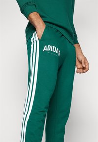 Zöld Adidas melegítőnadrág fehér oldalcsíkokkal és logó részletekkel. Puha anyagból készült, rugalmas derékkal. Oldalzsebekkel rendelkezik.
