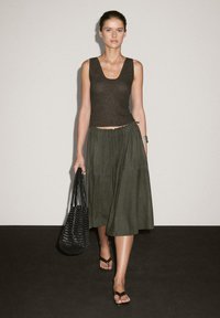 Modelen går selvsikkert iført en mørk strikket tanktop og en løsthængende olivengrøn nederdel, mens hun holder en vævet sort håndtaske. Enkle, neutrale baggrunde.