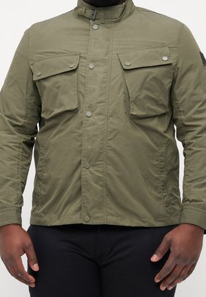 Hombre que lleva una chaqueta verde oliva abotonada con dos bolsillos de solapa en el pecho y pantalones negros sobre un fondo liso.