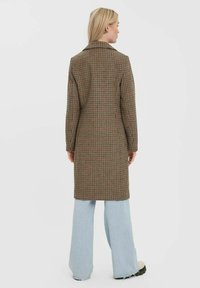 Hnědý károvaný dlouhý kabát s klopovým límcem, který má strukturovaný střih, zobrazený přes světle modré široké džíny.