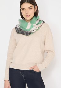 Maglione beige a maglia con scollo rotondo; abbinato a una sciarpa infinity verde, nera e bianca con motivi astratti e texture morbida.