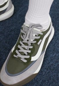 Zelena i siva tenisica sa teksturiranim gornjim dijelom, okruglim pertlama, bijelim detaljima i mekanim potplatom. Smještena na plavoj pozadini.