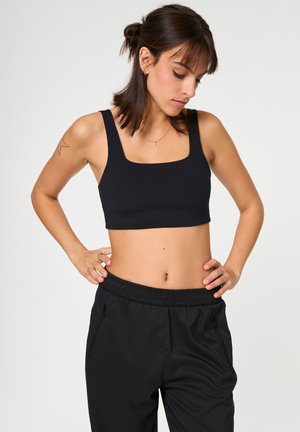 Νεαρή γυναίκα με σκούρα μαλλιά σε κότσο φοράει μαύρο κοντό τοπ και μαύρο παντελόνι με ελαστική μέση, κοιτάζοντας προς τα κάτω με τα χέρια στη μέση.