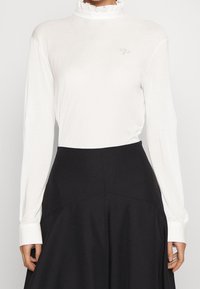 Vit långärmad topp med rynkad hög hals och en diskret logotyp, tillsammans med en svart utsvängd kjol med sömdetaljer.