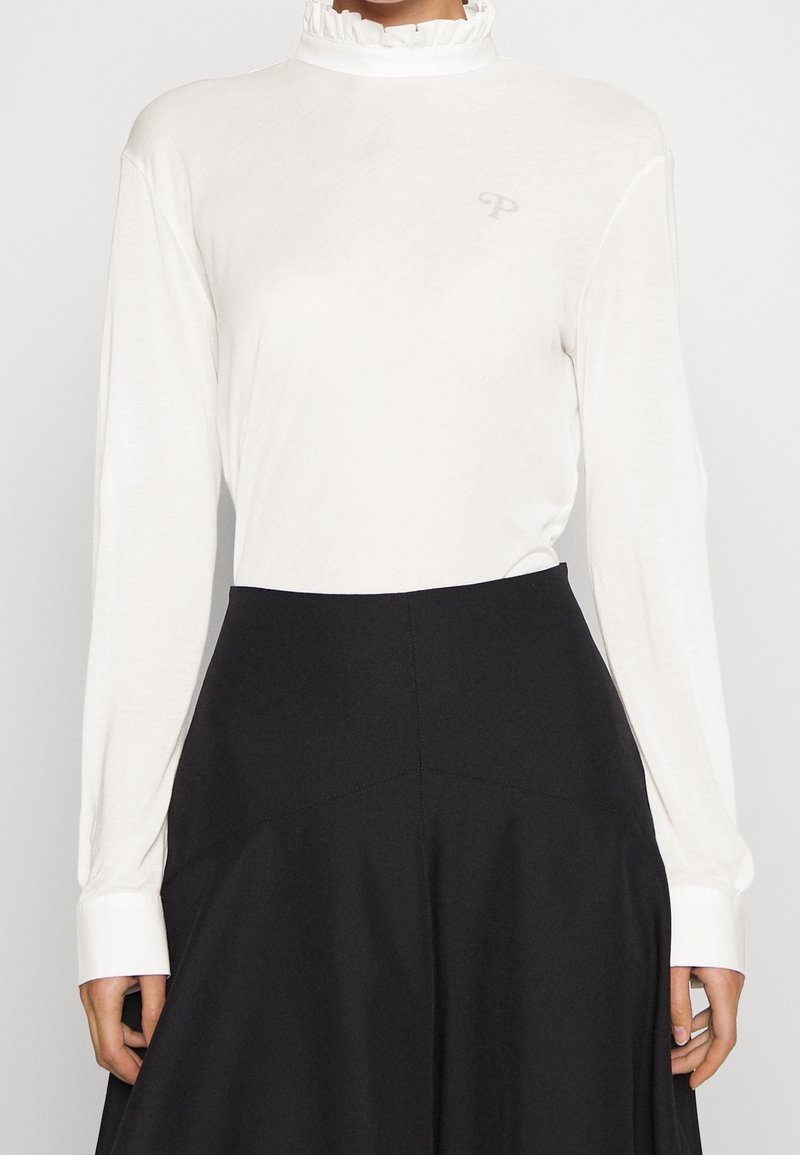 Vit långärmad topp med rynkad hög hals och en diskret logotyp, tillsammans med en svart utsvängd kjol med sömdetaljer.