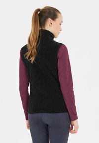 Sort fleecevest med høj krave, båret over en langærmet bordeaux top. Har et tætsiddende design og blød tekstur.