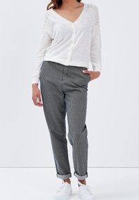 Vit långärmad kofta med spetsdetaljer vid axlarna, kombinerad med svart-vitrandiga byxor och vita sneakers.