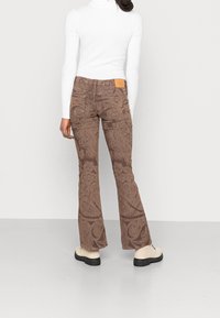 Bruna mönstrade utsvängda jeans med ett paisleymönster, med en beige läderlapp på baksidan, kombinerat med en vit långärmad topp och ljusa skor.