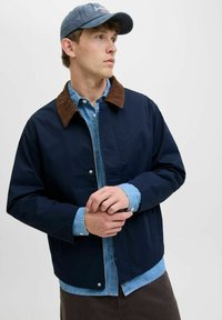 Marinejakke med krave i brunt corduroy, lynlåslukning og trykknapper, lagd over en lyseblå denimskjorte.