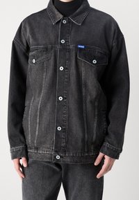 Chaqueta vaquera negra con cierre de botones, dos bolsillos en el pecho y una parche de logo azul. Presenta un cuello clásico y un ajuste holgado.