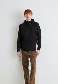 Felpa nera con zip e collo alto, tasche frontali e una texture liscia, abbinata a pantaloni marroni dritti. Design semplice e moderno.