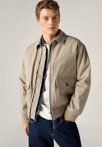 Νέος άνδρας που φοράει μπεζ ζακέτα με φερμουάρ πάνω από τζιν πουκάμισο και λευκό μπλουζάκι, με σκούρο μπλε τζιν, στέκεται μπροστά σε απλό φόντο.