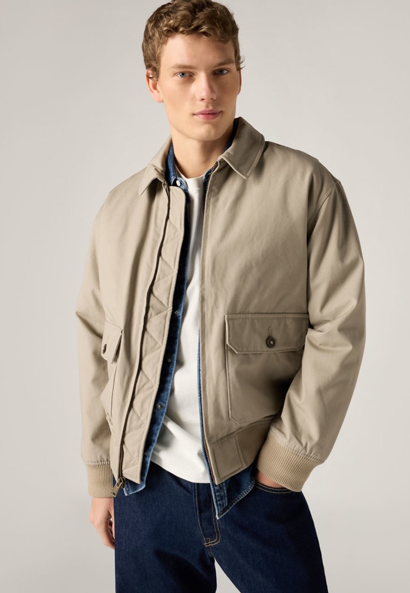 Giovane uomo con giacca beige con zip sopra una camicia di jeans e una maglietta bianca, indossa jeans blu scuro, in piedi davanti a uno sfondo semplice.