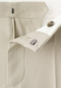 Tissu beige à texture lisse, avec une fermeture éclair et un bouton. Les éléments métalliques remarquables incluent un accent métallique et des détails de couture.