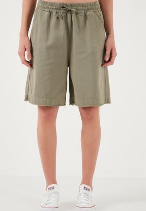 Osoba nosiaca olivovo zelené šortky na stiahnutie s ošúchanými okrajmi a bielymi nízkymi teniskami, stojaca pred jednoduchým svetlým pozadím.