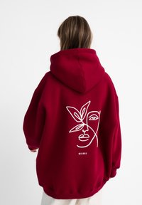 Rode oversized hoodie met een witte lijntekening van een gezicht en bladeren op de achterkant. Heeft een grote capuchon en geribde boorden.