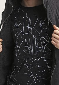 T-shirt nera con un design grafico bianco simile a schizzi che presenta il testo "BLACK KAVIER". Il tessuto sembra morbido con una vestibilità standard.