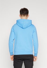 Felpa blu di cotone con tasca a canguro, polsi a coste con accenti rossi e bianchi, vista posteriore, texture morbida, design casual.