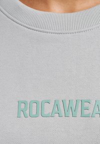 Svetlosivá mikina s rebrovaným výstrihom a embosovaným mätovo zeleným logom "ROC-A-WEAR". Materiál vyzerá hladko s pletenou textúrou.