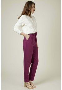 Pull en maille blanc avec un motif en câble, associé à un pantalon violet taille haute avec une ceinture et un devant plissé. Sandales à talons beiges.