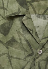 Tela verde con estampado que presenta formas abstractas, con un cuello con muesca, detalle de botones y una textura suave.