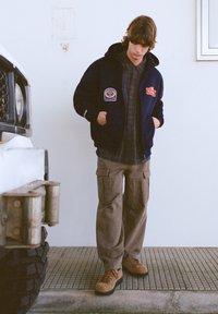 Námornícka kapucňová bunda s nášivkami, pod ňou kockovaná košeľa, béžové cargo nohavice a hnedé šnurovacie topánky. Textúrovaný povrch a pohodlný strih.