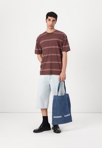 Brown striped t-shirt, light denim shorts, blue tote bag with white logo, black shoes, standing against a neutral background.