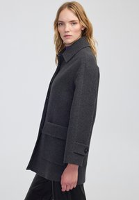 Cappotto in misto lana grigio con silhouette strutturata, ampio colletto, tasche frontali e dettagli sui polsini con bottoni. Tessuto liscio, design di lunghezza media.