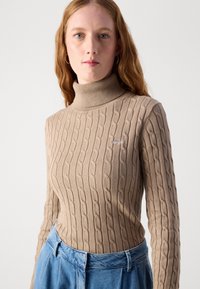Pull à col roulé beige à texture côtelée avec une silhouette ajustée, présentant un motif en tricot torsadé et un petit logo sur la poitrine.