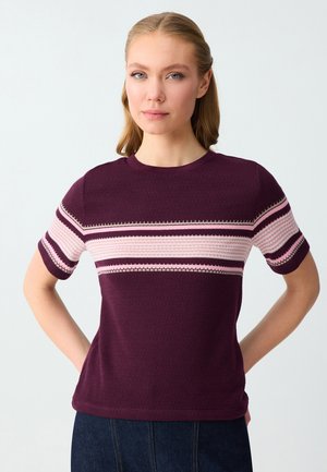 Donna con capelli biondi che indossa una maglietta viola a maniche corte con strisce orizzontali rosa e bianche, con le mani riposanti dietro la schiena.