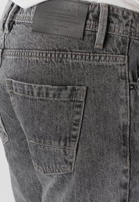 Nahaufnahme der Gesäßtasche einer grauen Jeans mit sichtbaren Nähten und einem schwarzen Lederetikett über dem Bund.