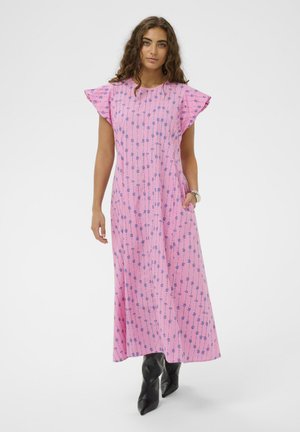 Femme portant une robe rose longueur cheville avec des rayures florales bleues et des manches volantes, assortie à des bottes noires pointues et un bracelet argenté.