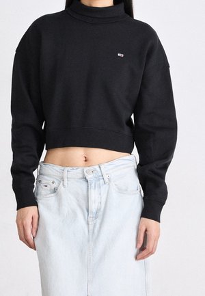 Persoon draagt een zwart cropped coltrui-sweatshirt gecombineerd met een lichtgewassen denim rok, waarbij de buik zichtbaar is en de handen ontspannen hangen.