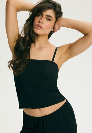 Kobieta z długimi ciemnymi włosami ubrana w czarny, bezrękawnik i dopasowane spodnie, pozująca z uniesionymi ramionami i rękami blisko głowy.