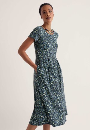 Femme debout, les mains dans les poches, portant une robe à fleurs bleu foncé à manches courtes, longueur genou, et un collier vert, tournée vers la gauche.