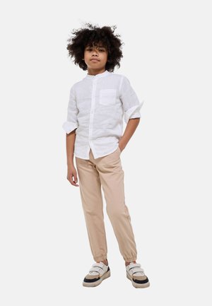 Garçon aux cheveux bouclés portant une chemise blanche à boutons, un pantalon de jogging beige et des baskets blanches à scratch, debout avec une main dans la poche.