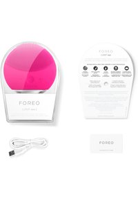 Dispositif de nettoyage facial en silicone rose compact avec surface texturée, câble de chargement USB, carte de garantie et informations sur le produit dans un emballage transparent.