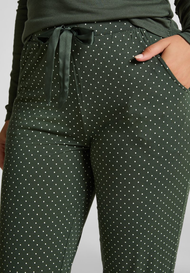 Groene broek met stippen in een zachte textuur, voorzien van een satijnen strikband en zakken. De stof heeft een subtiel geribbeld patroon.