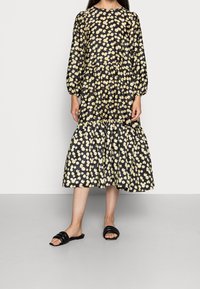 Schwarzes Kleid mit gelbem Blumenmuster, Puffärmeln, in der Taille gerafft und mit einem stufenförmigen Rock; kombiniert mit schwarzen Sandalen.