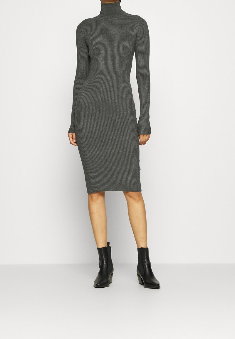 Femme portant une robe grise moulante à manches longues et col roulé côtelé, avec des bottines noires, debout devant un fond blanc.