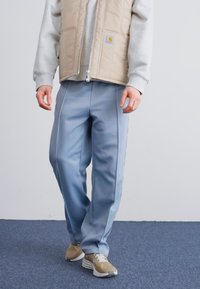 Pantaloni da jogging in poliestere blu chiaro con cuciture laterali, indossati con un gilet trapuntato beige e una felpa grigia; abbinati a sneaker chiare.
