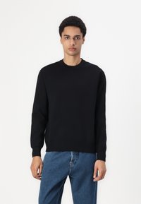 Pulover tricotat negru cu guler rotund, mâneci lungi și manșete și tiv cu nervuri. Asortat cu blugi din denim albăstrui.