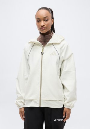 Žena v bielej bunde na zips cez hnedý top a čierne nohavice s logom „TERREX“, stojaca pred jednoduchým svetlým pozadím.