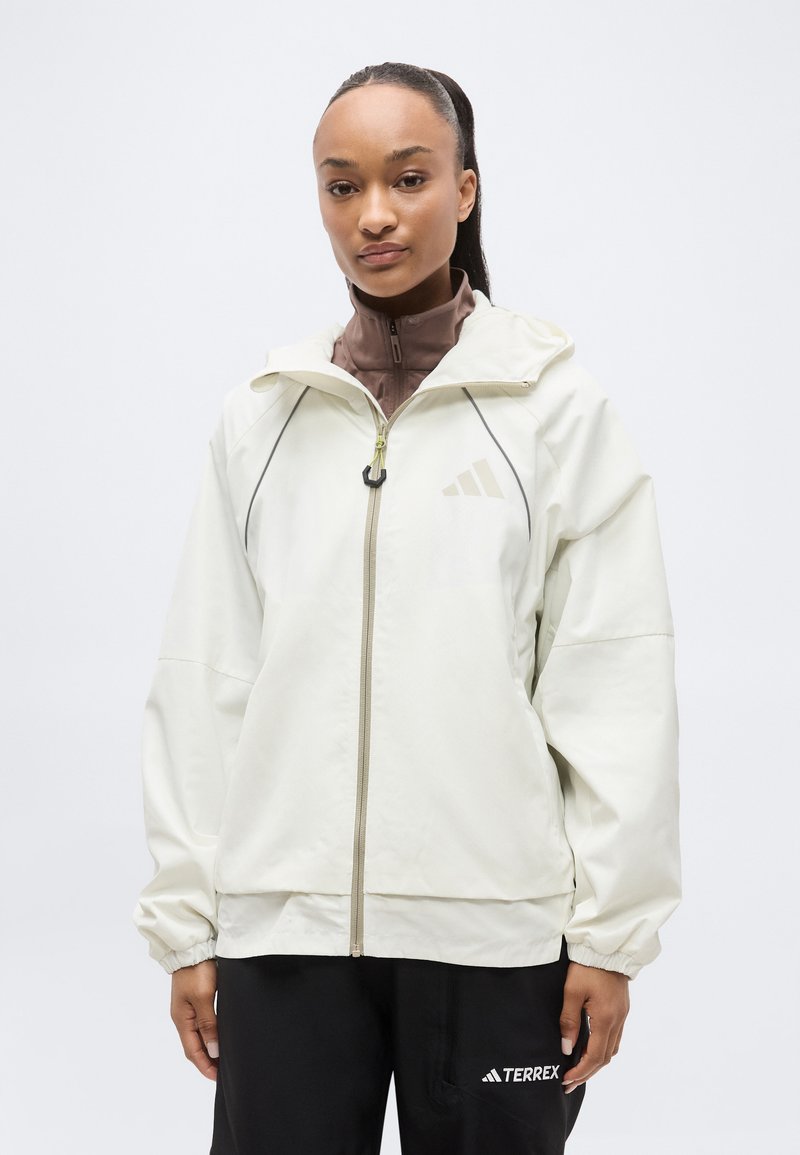 Femme portant une veste blanche zippée sur un haut marron et un pantalon noir avec le logo "TERREX", debout devant un fond clair uni.