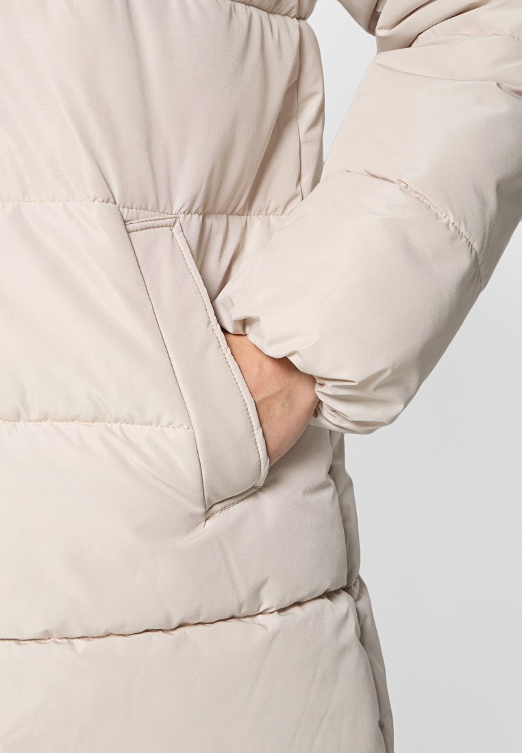 Chaqueta tipo puffer de color beige claro con un acabado texturizado y suave, puños elásticos y un bolsillo lateral que resalta el patrón acolchado.