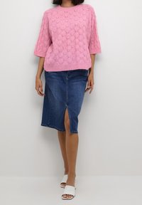 Rosa gestrickter Pullover mit strukturiertem Muster, kombiniert mit einem blauen Denim-Bleistiftrock mit einem Schlitz vorne. Dazu weiße Mules mit offenem Zeh.