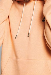 Persikfärgad hoodie med en känguruficka, dragsko i huvan och detaljer i silver. Mjuk textur, rundade axlar och en logotypdetalj.