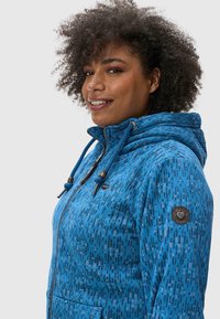 Hoodie bleu à motif avec fermeture éclair, capuche réglable à cordon, et deux poches avant ; possède un badge sur la manche gauche.