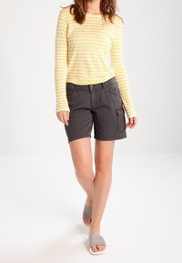 W paski, żółto-biała koszula z długim rękawem, zestawiona z szarymi szortami cargo z bocznymi kieszeniami, uzupełniona jasnoszarymi sandałami na wsuwkę.